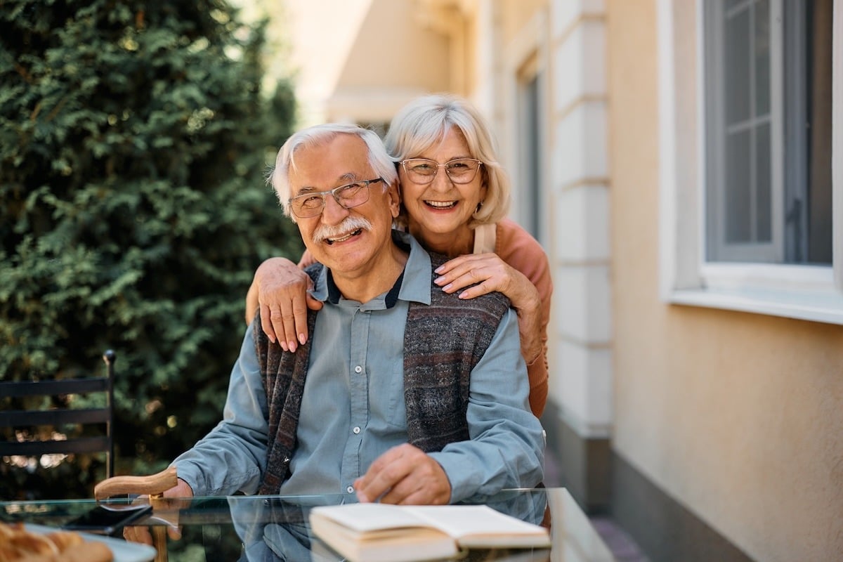 Elderly couple smiling outdoors at a glass table with an open book, surrounded by greenery and a building exterior.