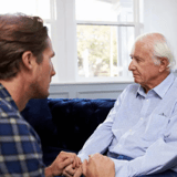 Two people sitting on a sofa, holding hands and facing each other in conversation.