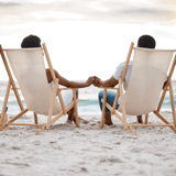 Two people holding hands while sitting on beach chairs facing the ocean.