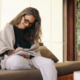 A woman with gray hair and glasses writing in a notebook, wearing a shawl and seated by a window.