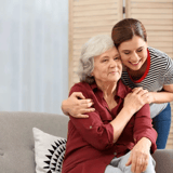 A young woman embraces an elderly woman sitting on a sofa.