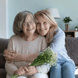 Two women sitting on a couch, one holding a bouquet, with a lamp and plant in the background.