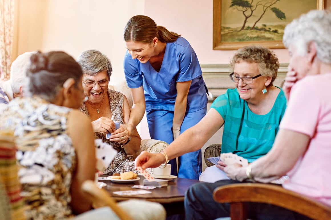 Group of assisted living residents playing cards game with nurse assisting