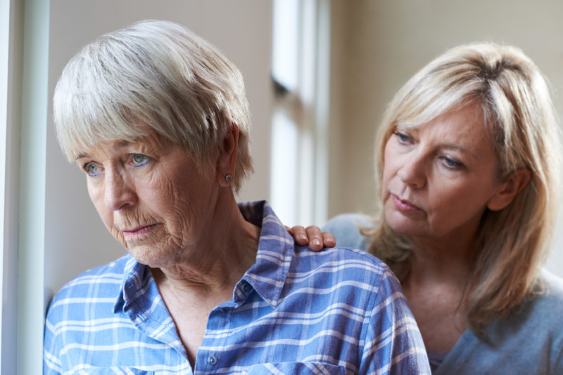 Senior woman looking out window while adult woman rests her hand on the senior's shoulder